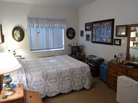 A cozy bedroom with a bed covered in a white and blue patterned quilt. There is a wooden nightstand with a lamp on the left side of the bed. The window has vertical blinds and a decorative valance. The walls are adorned with framed pictures and artwork. A wooden dresser with a mirror and various items on top is on the right side of the room. The room has a warm and homey atmosphere.
