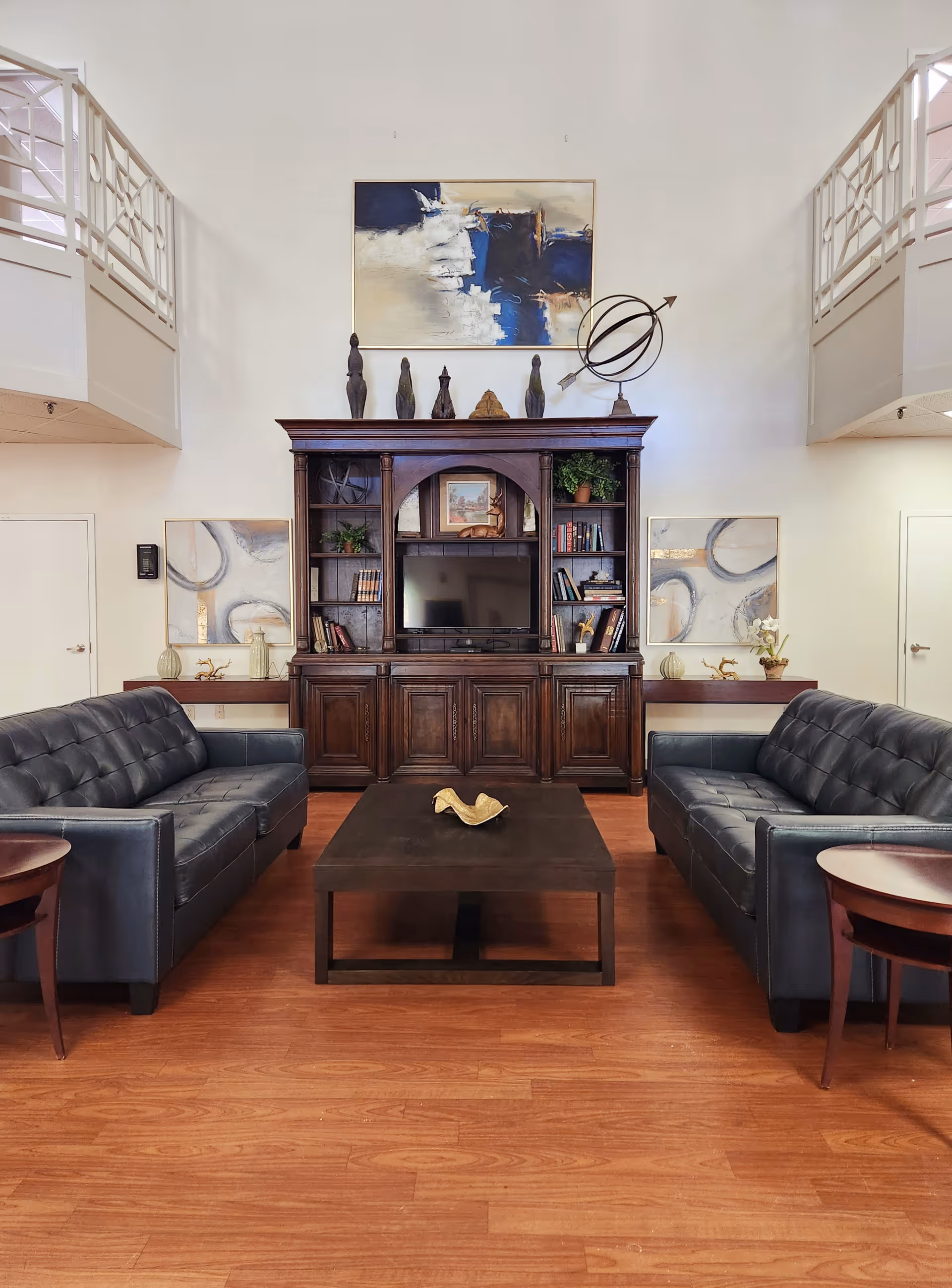 A cozy living room area with two dark leather sofas facing each other across a dark wooden coffee table. Behind the coffee table is a large wooden entertainment center with shelves holding books, plants, and decorative items, and a flat-screen TV in the center. Above the entertainment center is an abstract painting with blue, white, and beige tones, and a decorative armillary sphere. The room has wooden flooring and white walls with two doors on either side of the entertainment center.