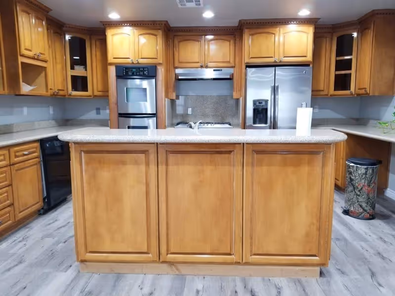 A modern kitchen with wooden cabinets and a large central island with a granite countertop. The kitchen features a stainless steel double oven, a stainless steel refrigerator with a water dispenser, a stovetop with a range hood, and a paper towel holder on the island. The floor is covered with light-colored wood laminate, and there is a camouflage-patterned trash can in the corner.