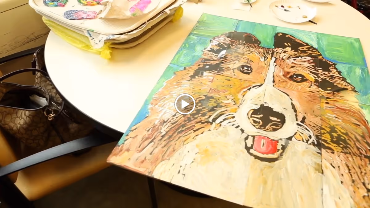 A painted artwork of a dog's face on a canvas placed on a round white table. Nearby are some art supplies and a brown handbag on a chair.