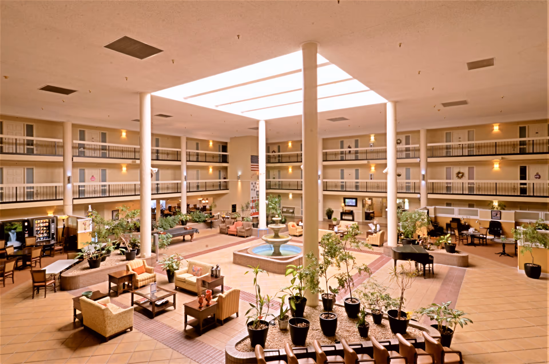 Spacious indoor atrium area with a central water fountain surrounded by potted plants and comfortable seating arrangements including armchairs and sofas. The atrium is illuminated by a large skylight overhead and features multiple floors with doors and railings overlooking the central space. Additional furnishings include a piano, a pool table, and vending machines along the walls.