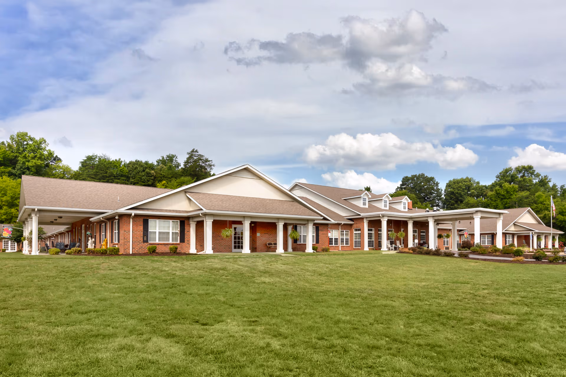 Front exterior of a single-story brick senior living building with a wide green lawn and covered porches under a partly cloudy sky.