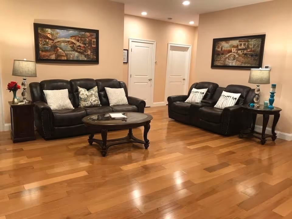 A living room with two dark brown leather sofas, each adorned with decorative pillows. Between the sofas is a round wooden coffee table with a book on it. On either side of the sofas are wooden side tables with lamps and decorative items. The walls are painted beige and feature framed paintings. The floor is made of polished wood, and there are two white doors in the background.