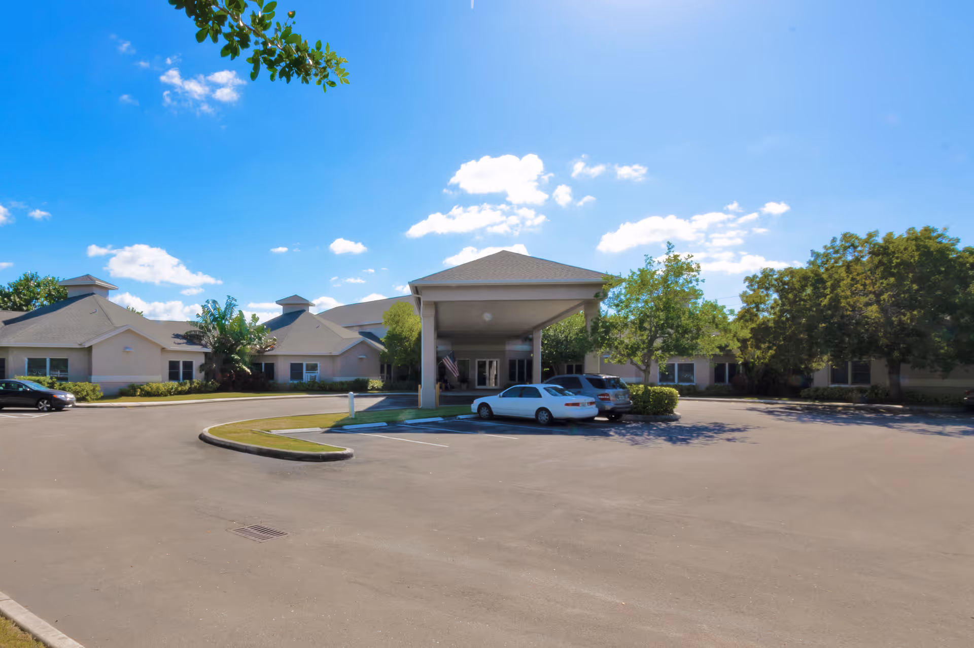 Front exterior of a single-story senior living facility with a covered entrance, parked cars, and landscaped grounds under a blue sky.