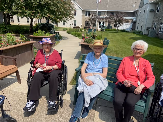 Three elderly women sitting outdoors on a sunny day at a senior living facility. One woman is in a wheelchair wearing a pink hat and red top, the middle woman is sitting on a green bench wearing a straw hat and blue shirt, and the third woman is also seated on the bench wearing glasses and a red jacket. Behind them is a garden area with raised flower beds and a pathway leading to a large building.