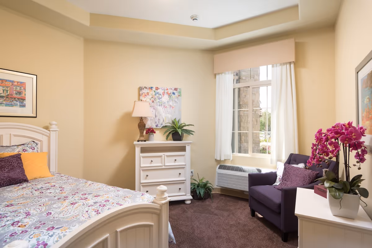 A cozy bedroom with a white wooden bed featuring a floral patterned bedspread and colorful pillows. There is a white dresser with a lamp, a small plant, and a colorful painting above it. A purple armchair with a decorative pillow is placed near a window with white curtains. Another white piece of furniture holds a vibrant pink orchid plant. The walls are painted beige and the carpet is brown.
