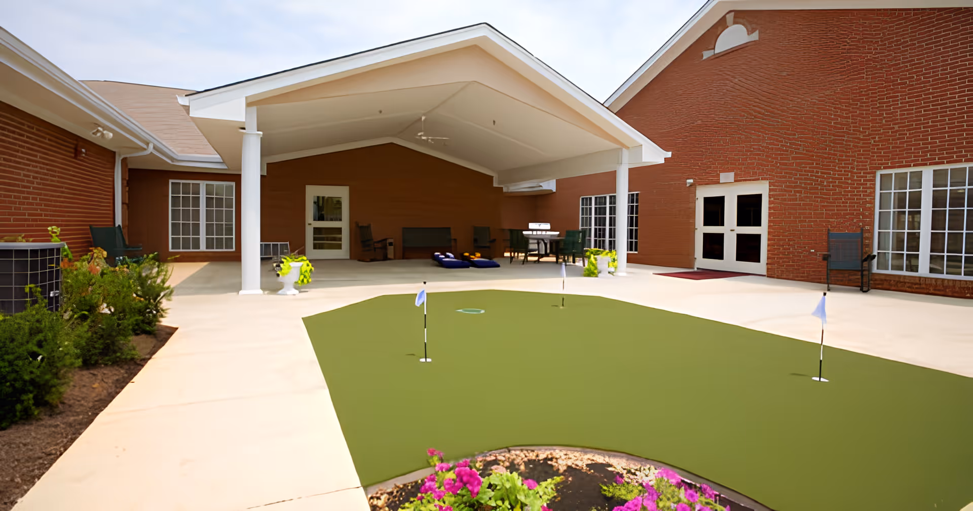 Outdoor courtyard area of a senior living facility with a small putting green, potted plants, patio furniture, and a covered seating area attached to a red brick building.