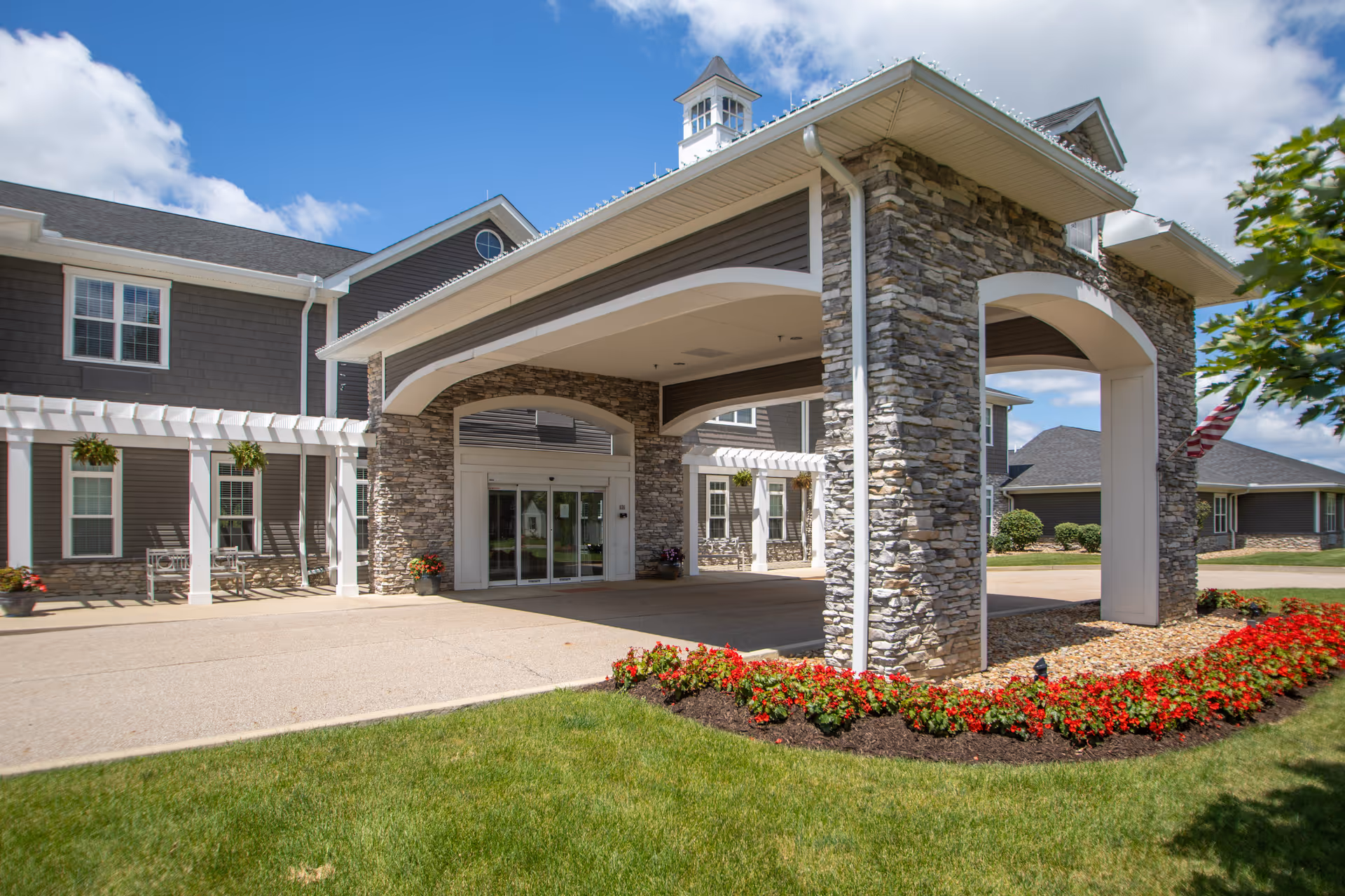 Front entrance of a senior living facility with a covered stone porte-cochère, glass doors, and landscaped flowerbeds.