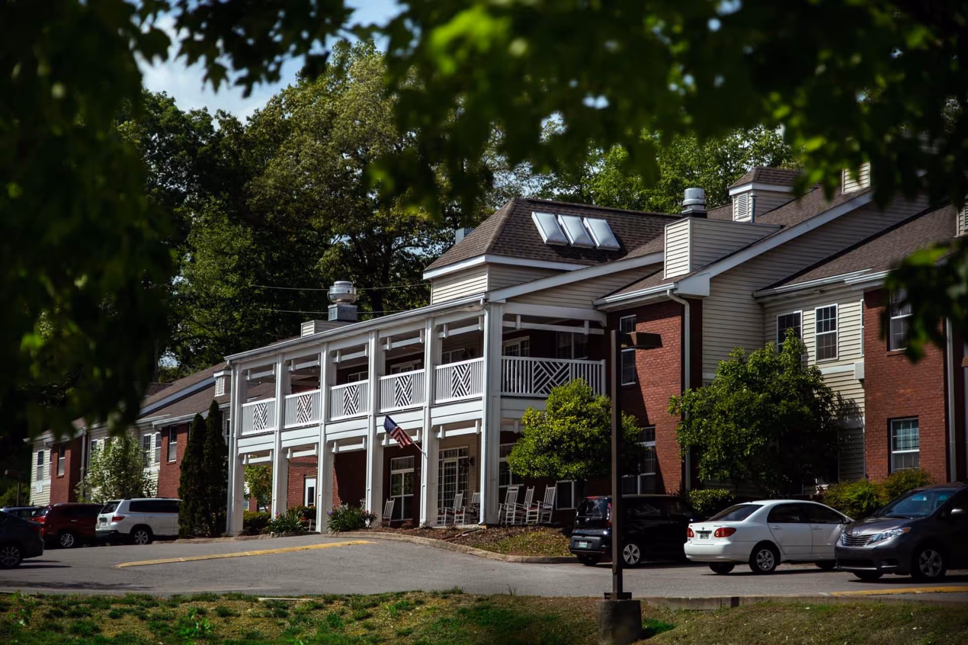 Exterior view of a two-story senior living facility building with a covered porch featuring white railings and rocking chairs. Several cars are parked in front of the building, which is surrounded by trees and greenery under a clear sky.