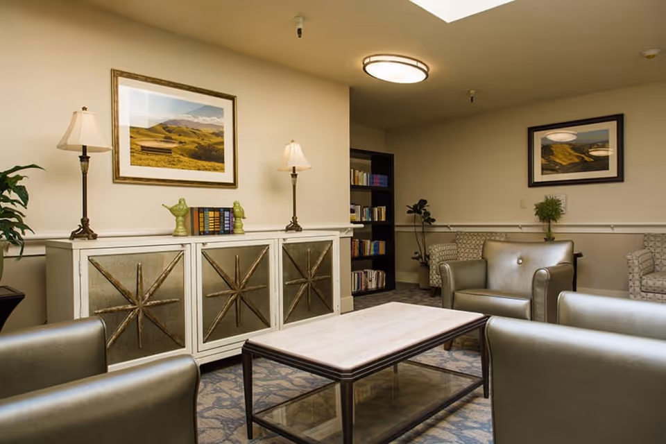 A cozy living room area with beige walls and carpeted floor. The room features a white cabinet with decorative star designs, two table lamps, and a few books and green bird figurines on top. There are several armchairs arranged around a rectangular coffee table with a glass shelf underneath. The walls are adorned with framed landscape photographs, and a bookshelf filled with books is visible in the corner.