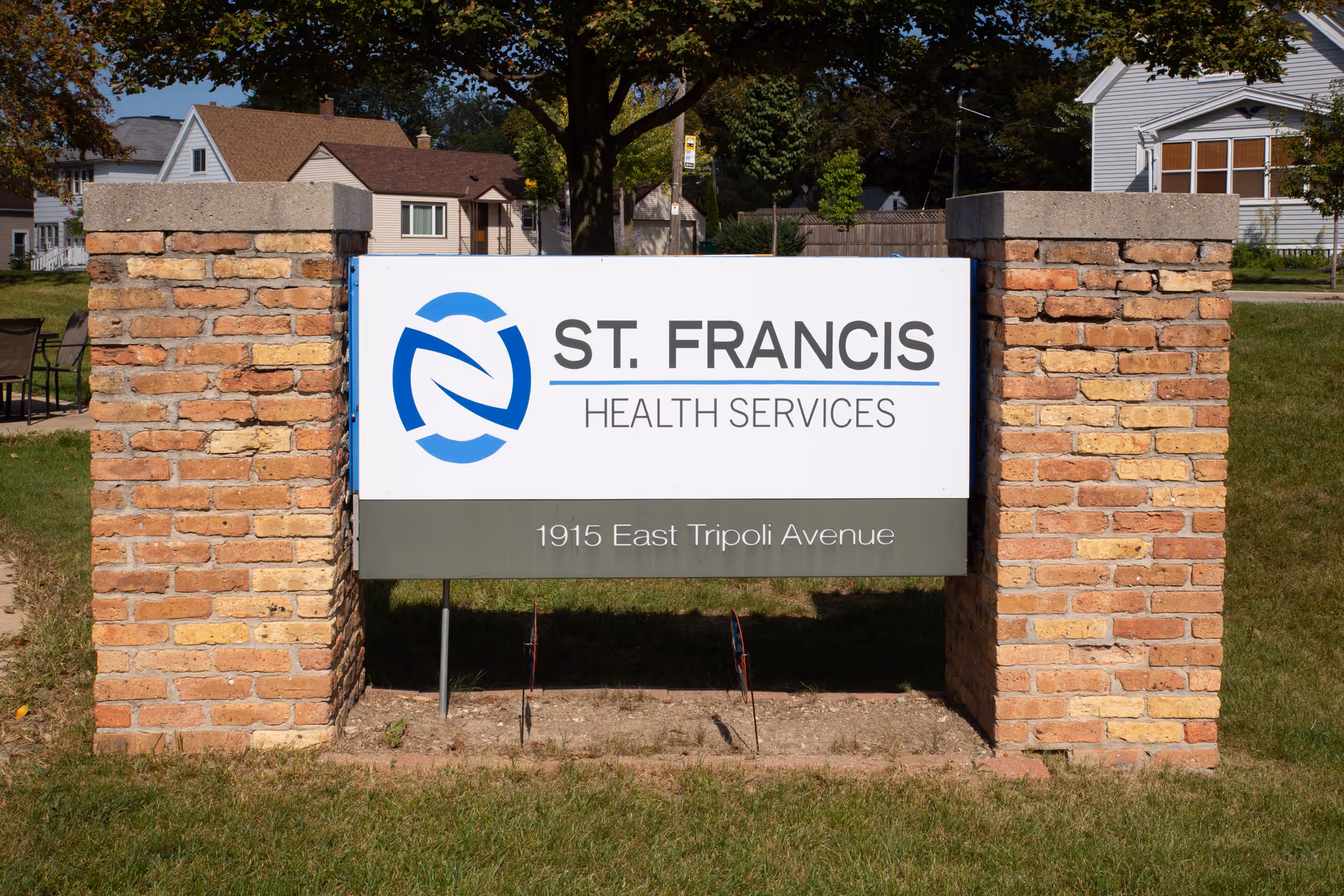 A sign for St. Francis Health Services mounted between two brick pillars on a grassy area with houses and trees in the background. The sign includes the address 1915 East Tripoli Avenue.