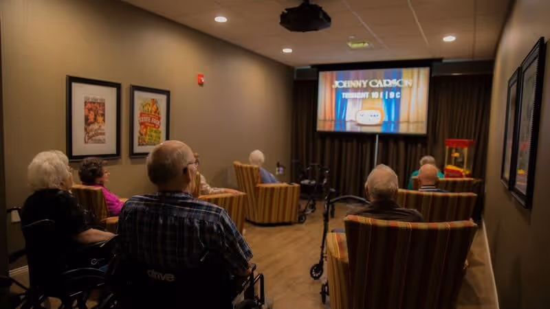 A group of elderly people seated in a small theater room watching a screen showing a Johnny Carson program. Some are in wheelchairs and others in striped armchairs. The room has framed pictures on the walls and a popcorn machine in the corner.