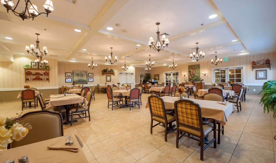 A spacious senior living dining room with multiple tables covered in beige tablecloths, each set with napkins and silverware. The room features elegant chandeliers hanging from a coffered ceiling, framed artwork on the walls, and a warm, inviting atmosphere with beige tiled floors and comfortable chairs.