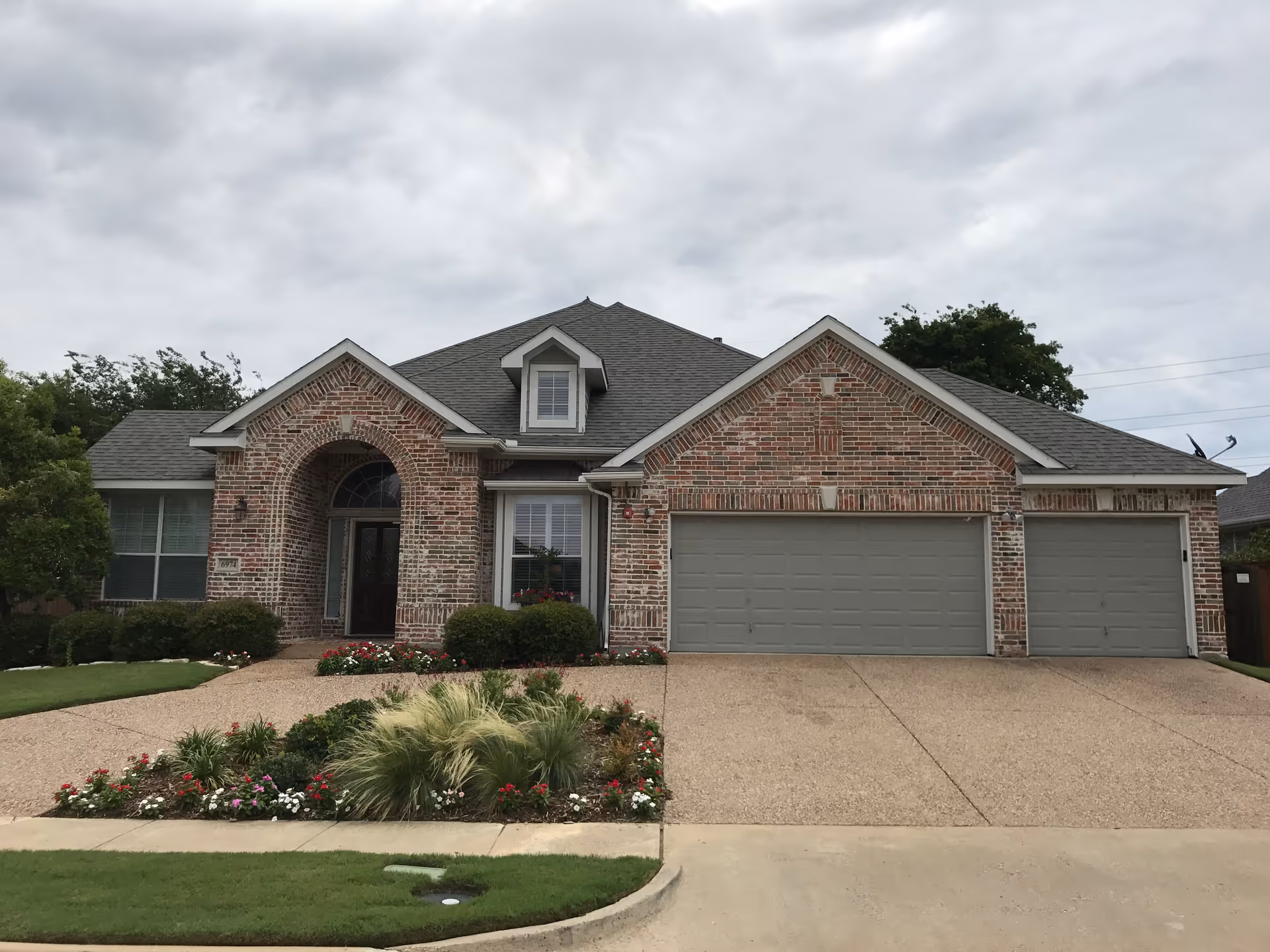 Front exterior view of a single-story brick house with a three-car garage, a small garden with flowers and shrubs, and a cloudy sky above.