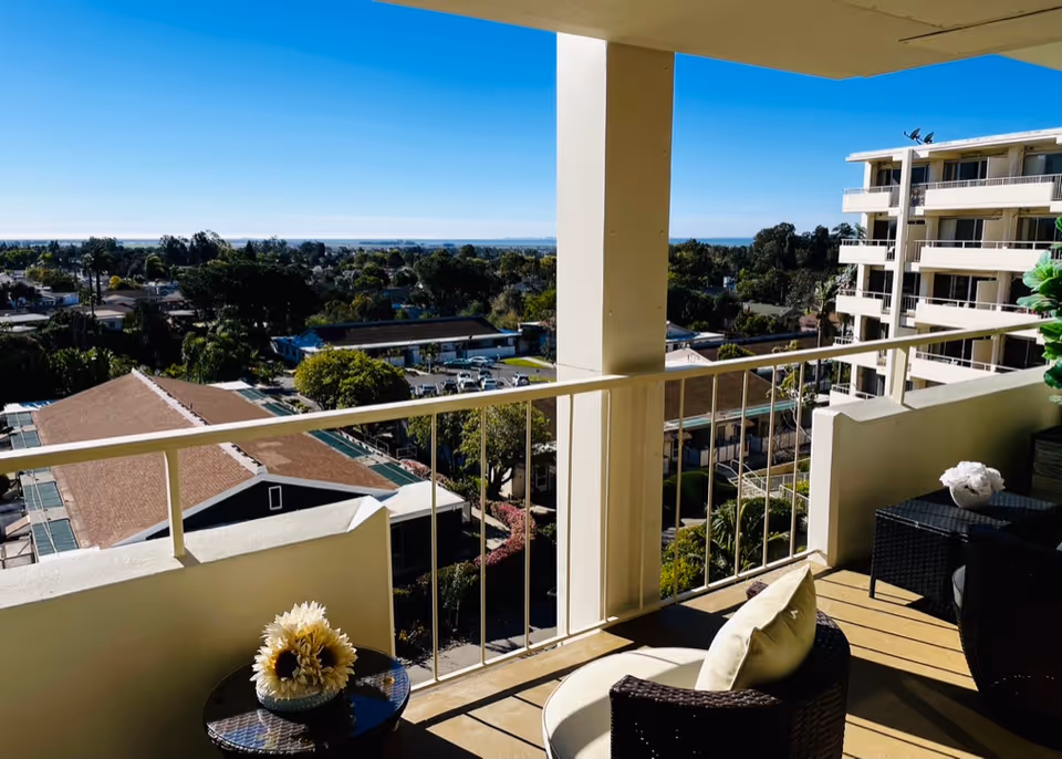 View from a balcony with outdoor seating including cushioned chairs and a small table with a flower arrangement, overlooking a residential area with trees and buildings under a clear blue sky.