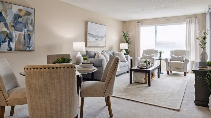A bright and cozy living room and dining area in a senior living facility. The living room features a light gray sofa with multiple cushions, two beige armchairs, a dark wooden coffee table with decorative items, and a large window with white curtains letting in natural light. The dining area has a round glass table surrounded by four beige upholstered chairs with nailhead trim. The walls are decorated with abstract paintings, and there are several potted plants adding greenery to the space.