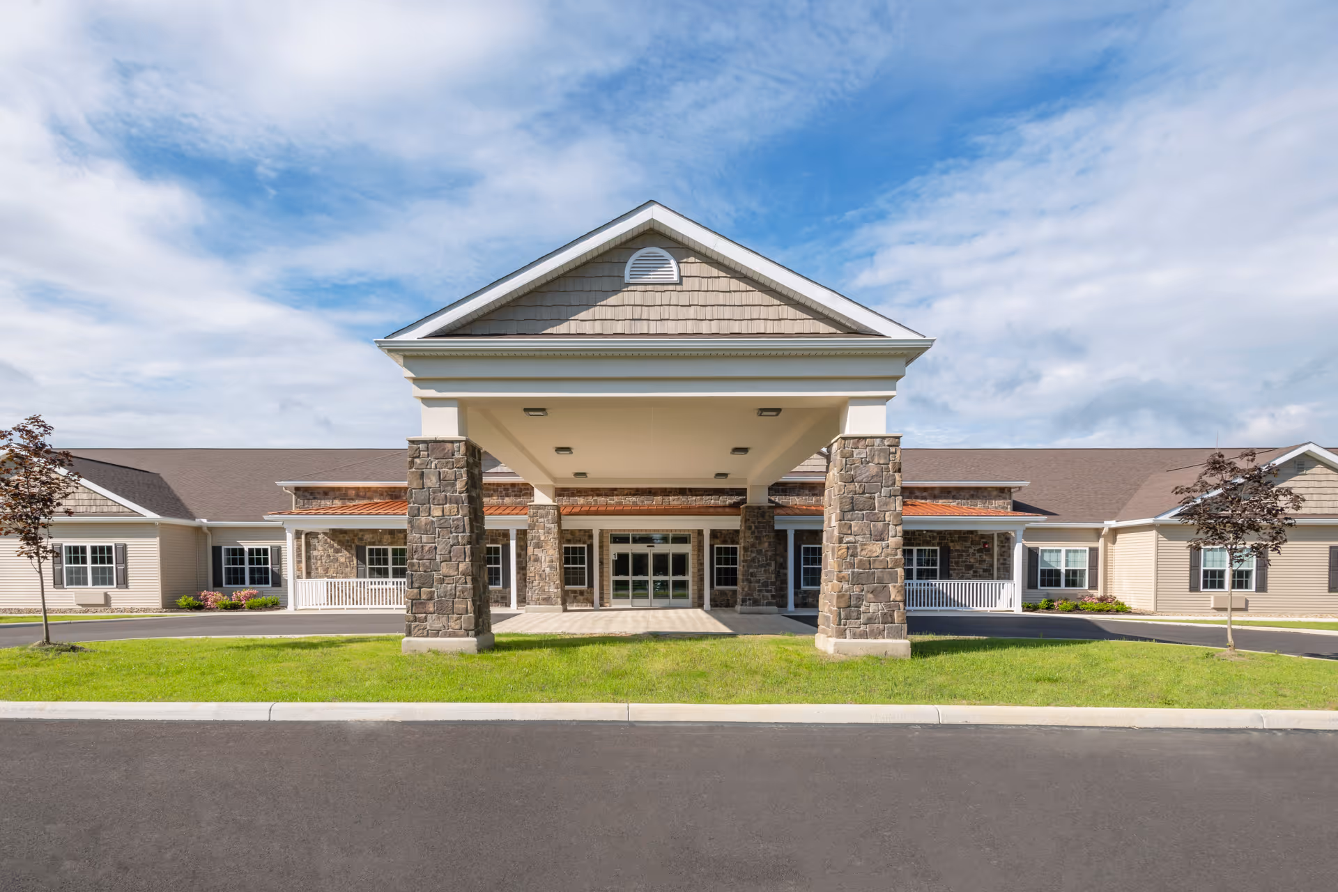 Front entrance of a single-story senior living facility with a covered porte-cochère supported by stone columns, a driveway and lawn in front.