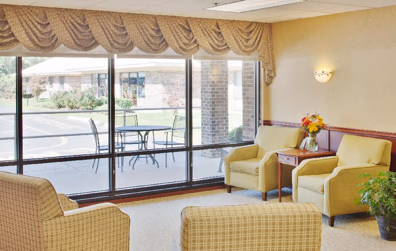 Sunlit seating area with upholstered armchairs and a small table holding flowers facing floor-to-ceiling windows and an outdoor patio.