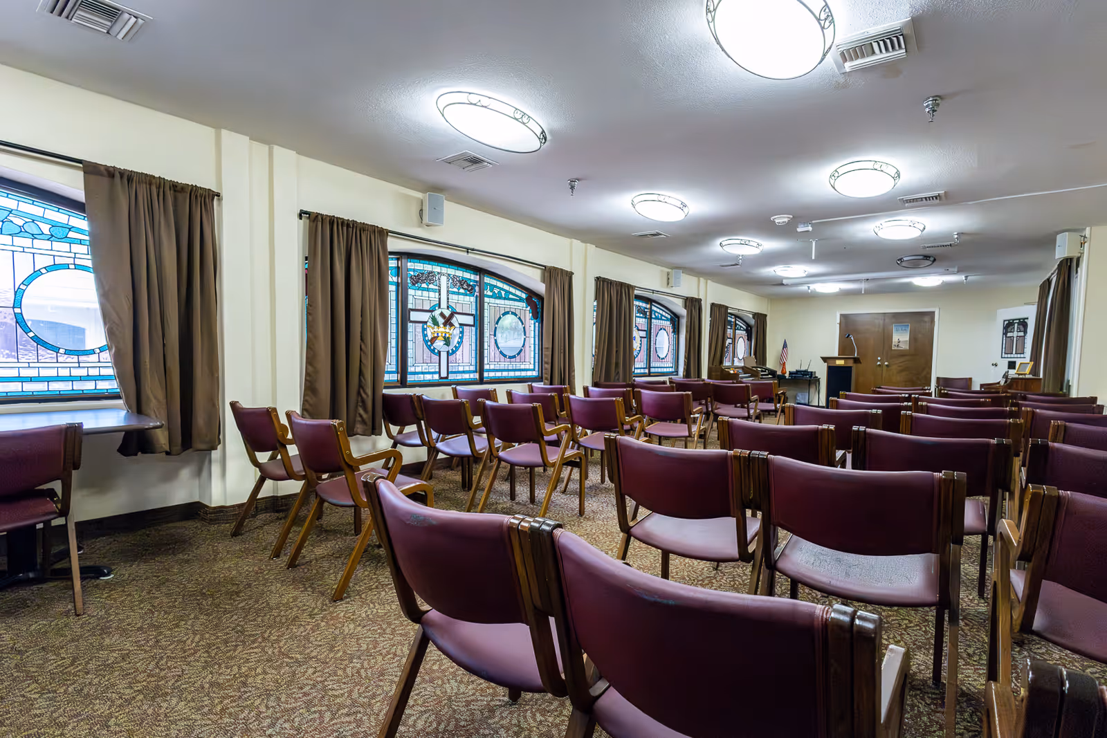 A room set up with multiple rows of purple chairs facing a podium and a piano at the front. The room has stained glass windows with brown curtains, carpeted floor, and circular ceiling lights.