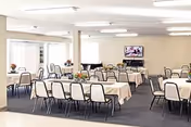 A spacious dining room with multiple rectangular tables covered with white tablecloths, each surrounded by chairs. The room has neutral-colored walls, carpeted flooring, and large windows allowing natural light. A television is mounted on the far wall, and there are flower arrangements on the tables.