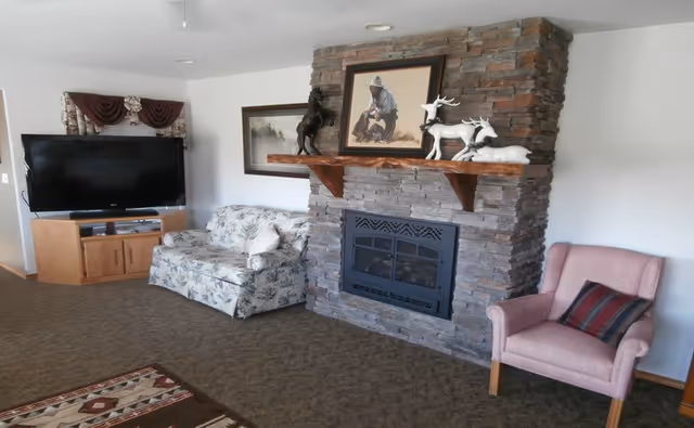 A cozy living room with a stone fireplace featuring a wooden mantel decorated with animal figurines and a framed painting. To the left of the fireplace is a floral patterned loveseat and a flat-screen TV on a wooden stand. To the right is a pink upholstered armchair with a striped cushion. The room has carpeted flooring and white walls.