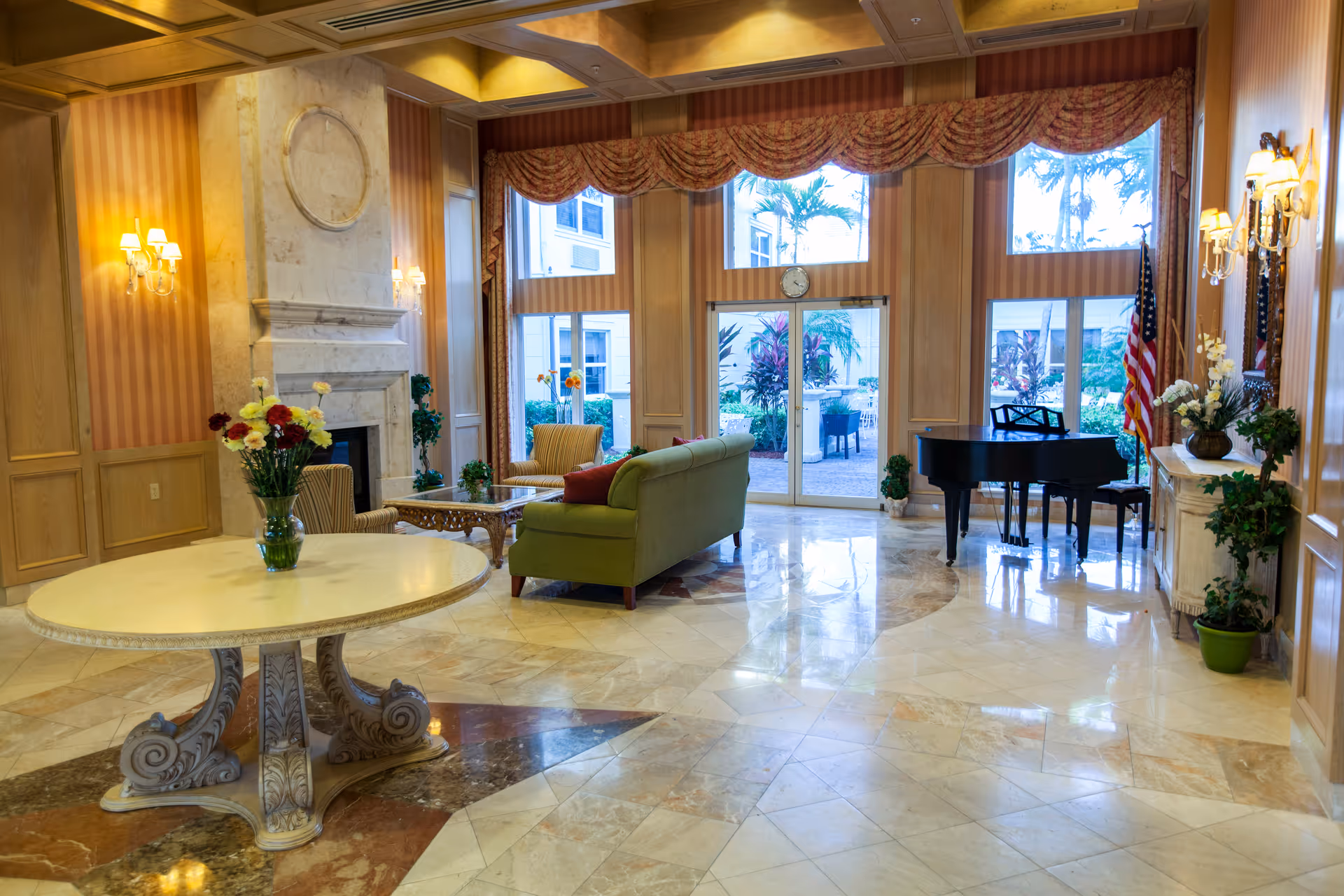 Sunlit lobby-style sitting area with marble floors, a round table with flowers, a fireplace, green sofa, and a grand piano by large windows.