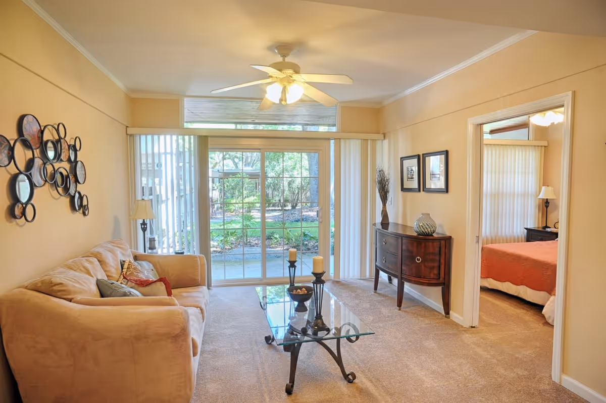 A cozy living room with a beige sofa adorned with colorful pillows, a glass coffee table with decorative candles and a bowl, a wooden sideboard with a vase, and sliding glass doors leading to a green outdoor area. The room has beige walls, carpeted floor, and a ceiling fan with lights. A doorway on the right leads to a bedroom with an orange bedspread and a lamp on a nightstand.