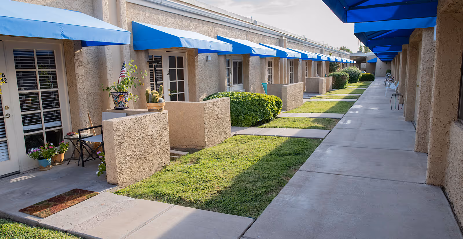 Row of ground-floor apartment entrances with blue awnings, small patios, potted plants, and a paved walkway beside grassy strips.