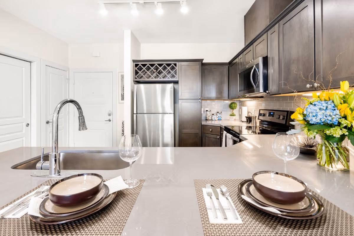 Modern kitchen with dark wood cabinets, stainless steel refrigerator and microwave, a sink with a curved faucet on a large countertop island set with two place settings including bowls, plates, utensils, and wine glasses. A vase with yellow and blue flowers is on the right side of the counter.