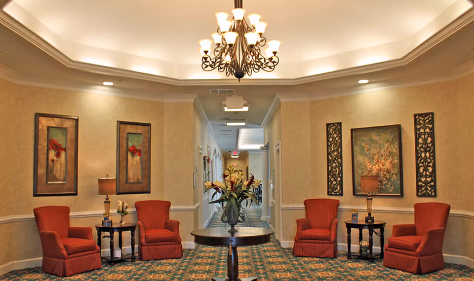 A cozy seating area in a senior living facility with four red upholstered armchairs arranged around two small dark wooden side tables with lamps. In the center is a round wooden table with a floral arrangement. The walls are decorated with framed floral artwork and decorative panels. The ceiling features a chandelier and recessed lighting, and a hallway extends in the background.