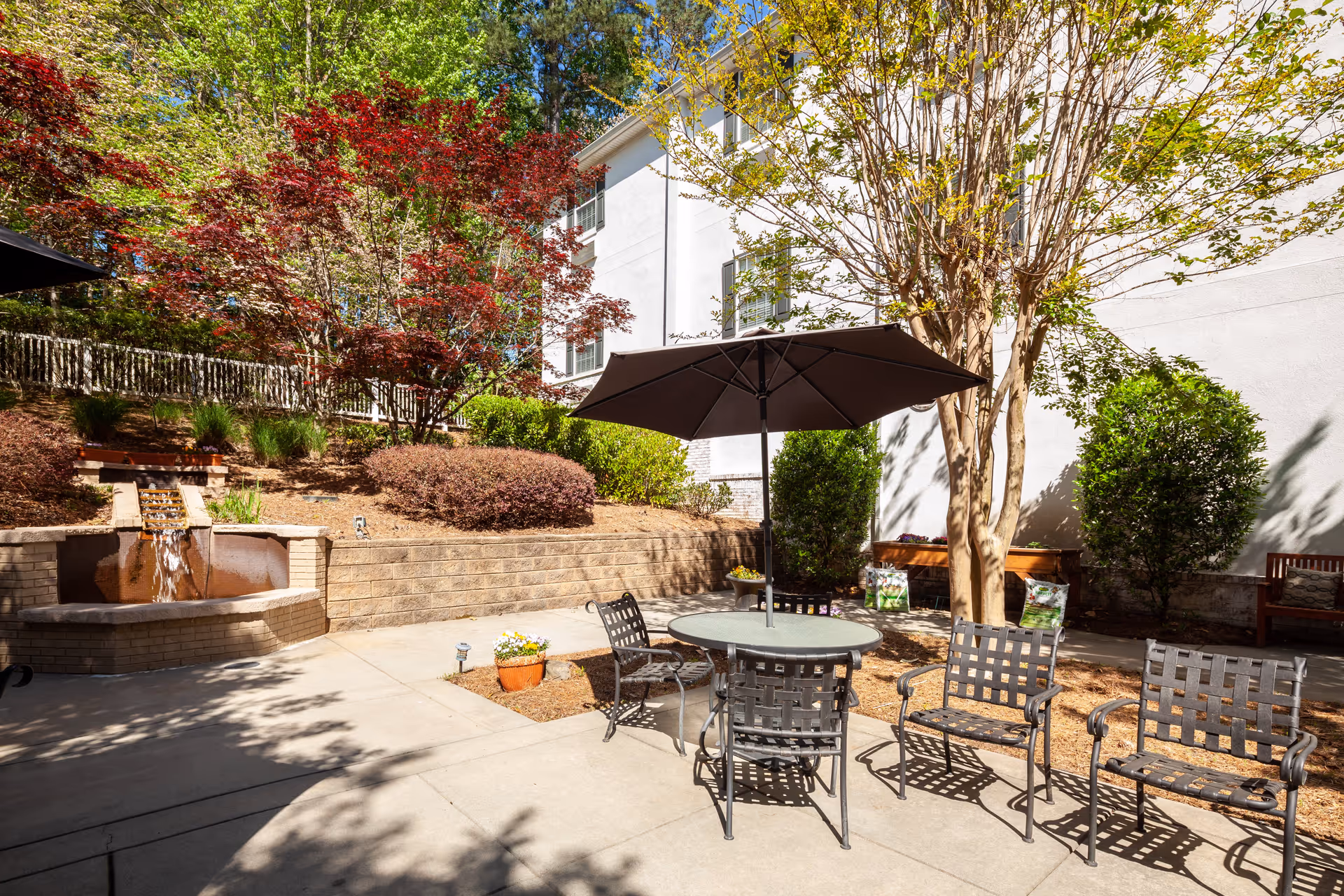 Outdoor patio area with a round table and four metal chairs under a large black umbrella. There is a small water fountain feature on the left side, surrounded by landscaped bushes and trees with red and green leaves. A white building wall is visible in the background with some benches and potted plants nearby.