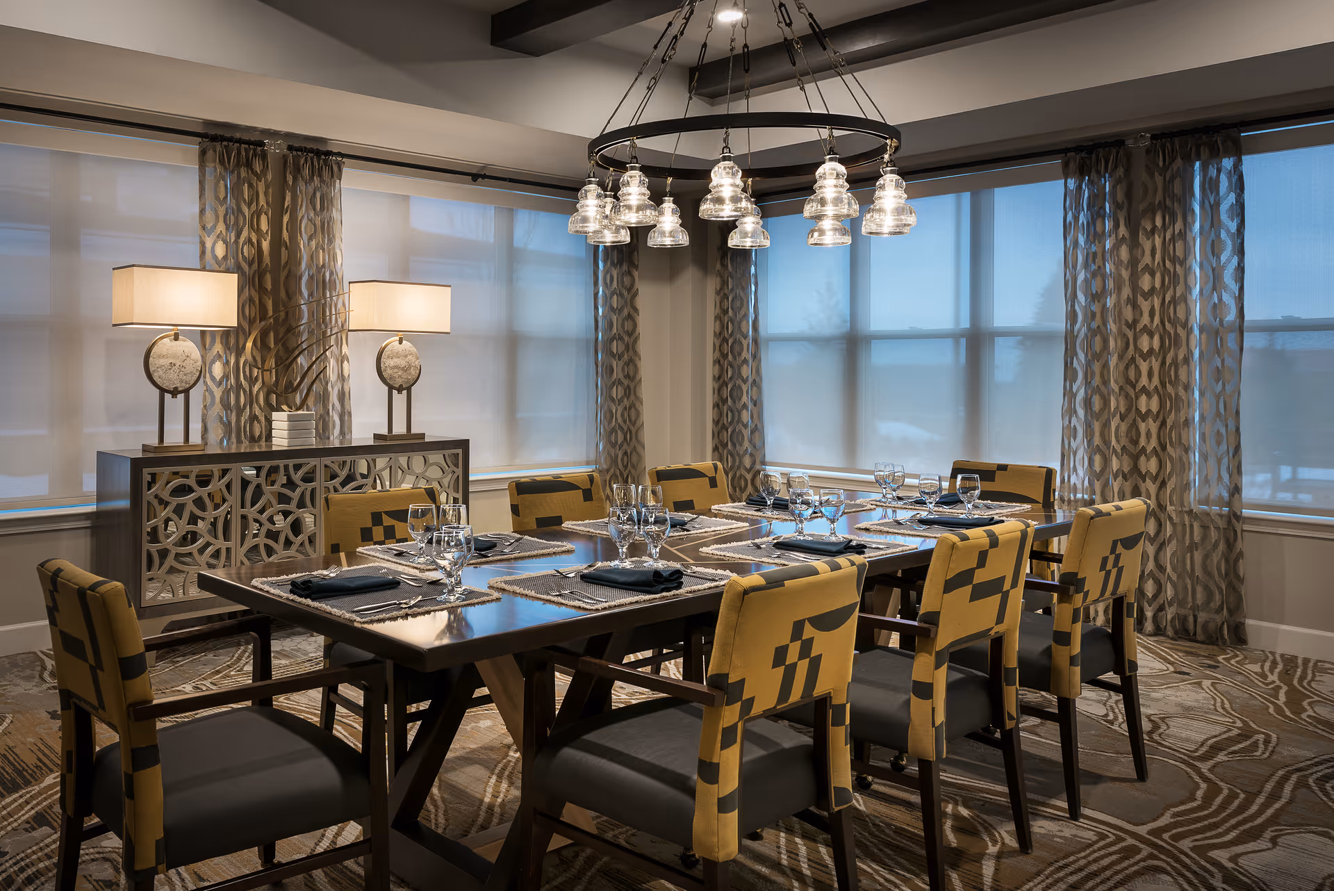 A well-lit dining room with a large rectangular wooden table set for eight people. The table has placemats, napkins, and glassware. Surrounding the table are eight upholstered chairs with a yellow and black geometric pattern. The room features large windows with patterned curtains and roller shades, a decorative sideboard with two table lamps, and a modern chandelier with multiple glass pendant lights hanging above the table.