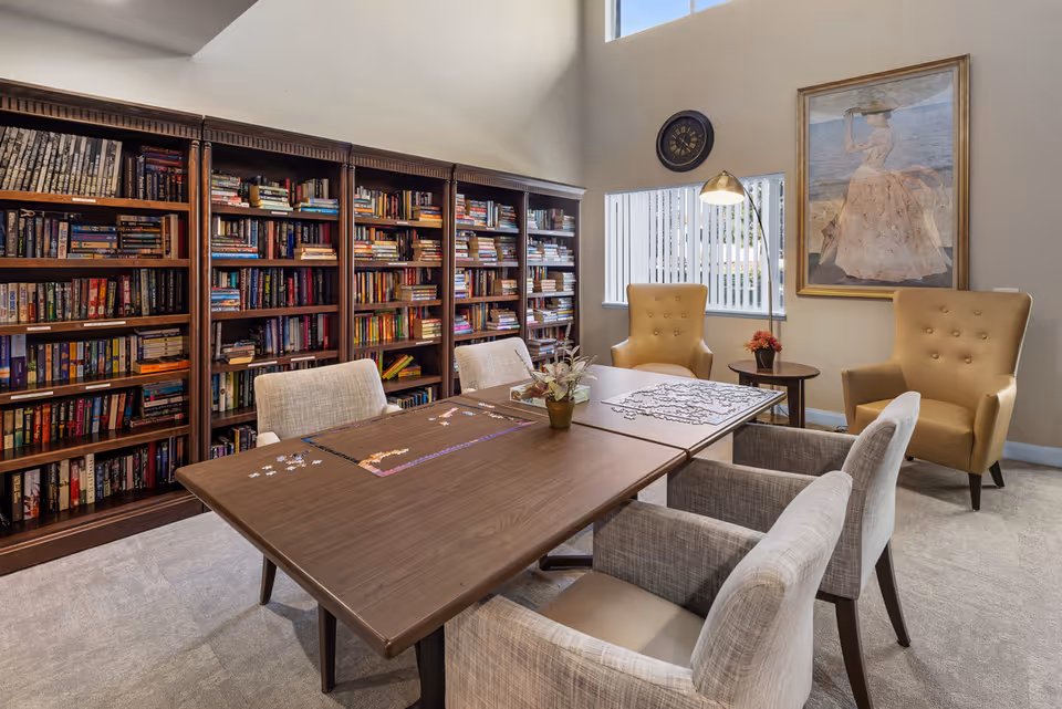 A cozy reading and activity room with a large wooden table in the center, partially covered with a puzzle in progress. Surrounding the table are four upholstered chairs and two tan armchairs near a window with vertical blinds. A tall wooden bookshelf filled with books lines one wall, and a framed painting of a woman in a hat is hung on another wall beneath a round clock. A floor lamp provides additional lighting.
