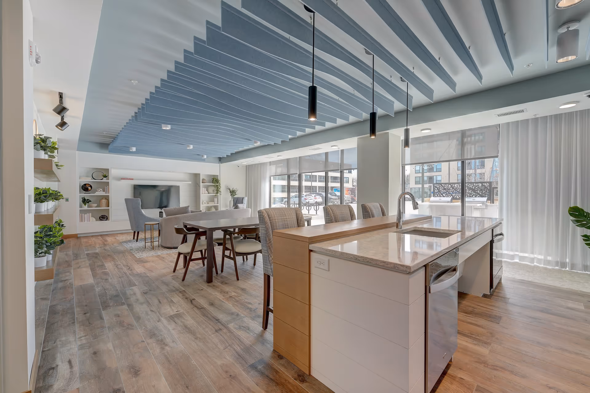 A modern, open-concept living space featuring a kitchen island with a sink and bar stools, a dining table with chairs, and a cozy seating area with armchairs and a TV. The room has large windows with sheer curtains allowing natural light to fill the space, wood flooring, and a unique blue ceiling design with hanging pendant lights.
