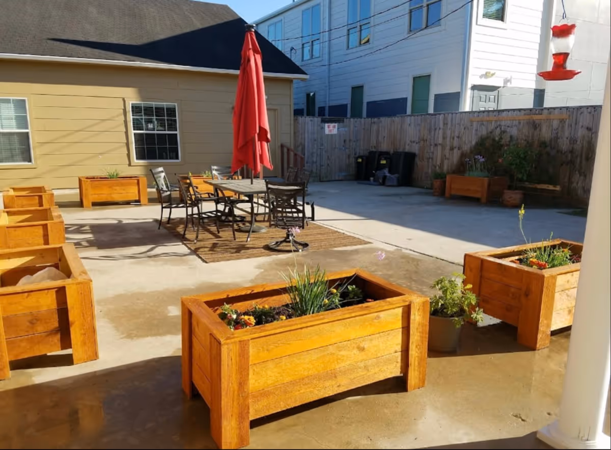 Sunlit outdoor courtyard with wooden planter boxes, a central dining table and red umbrella, and surrounding buildings and fence.