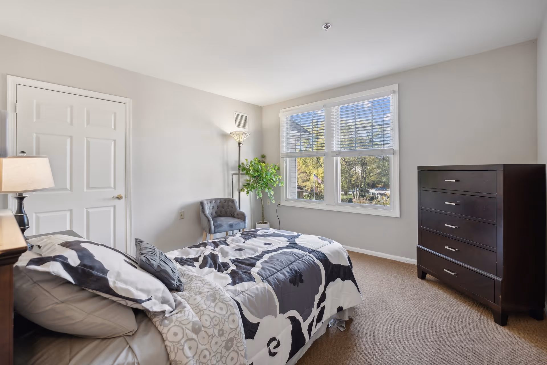A bright bedroom with a large window letting in natural light. The room features a bed with patterned bedding, a dark wooden dresser, a gray upholstered chair, a floor lamp, and a potted plant near the window. The walls are painted light gray and the carpet is beige.