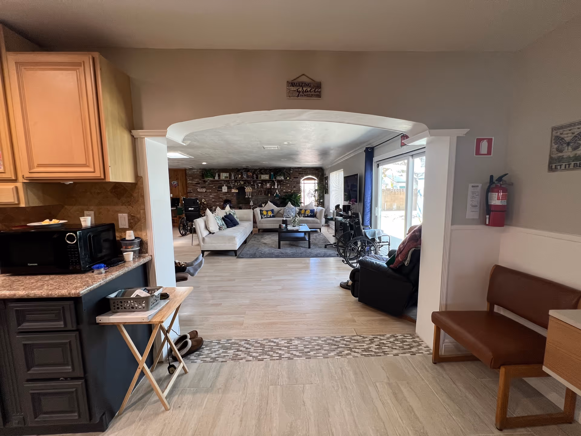 View from a kitchen area into a living room with light wood flooring, a white sofa set, a coffee table, and a TV mounted on the wall. The kitchen counter has a microwave and some cups. There is a fire extinguisher mounted on the wall to the right, a brown bench, and a wheelchair near the living room area. The living room has a brick accent wall with shelves and decorations.
