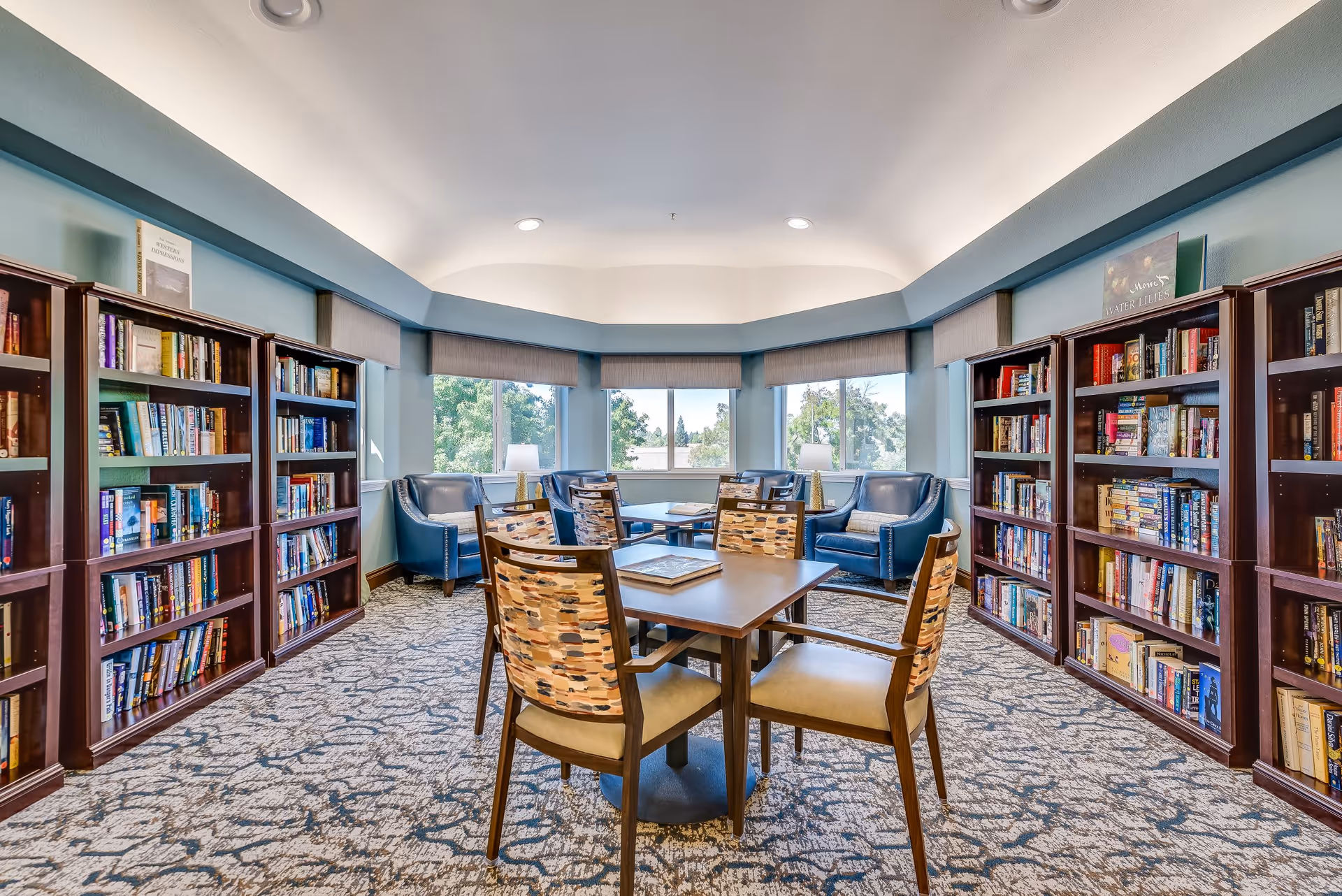 A bright and cozy library room with large windows letting in natural light. The room features multiple wooden bookshelves filled with books along the walls, a central table with four patterned chairs, and a seating area with four blue armchairs and two small lamps near the windows. The carpet has a patterned design, and the walls are painted a soft blue color.