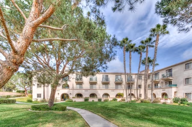 Exterior view of a three-story senior living facility building with arched windows and balconies, surrounded by green grass, tall palm trees, and other large trees under a partly cloudy sky.