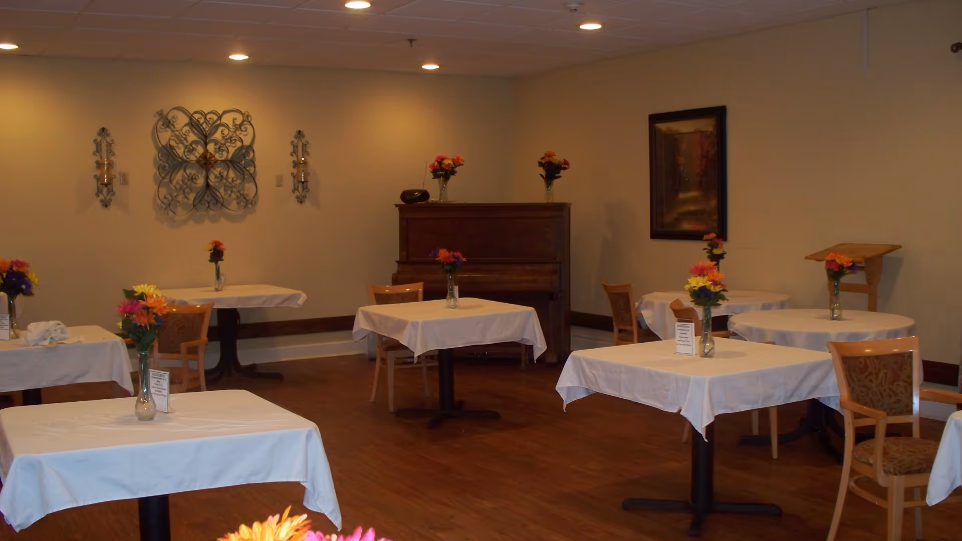 A dining room with several tables covered in white tablecloths, each adorned with a vase of colorful flowers. Wooden chairs are placed around the tables. The room has wooden flooring, beige walls, decorative wall hangings, a piano against the far wall, and a framed painting. The lighting is provided by recessed ceiling lights.