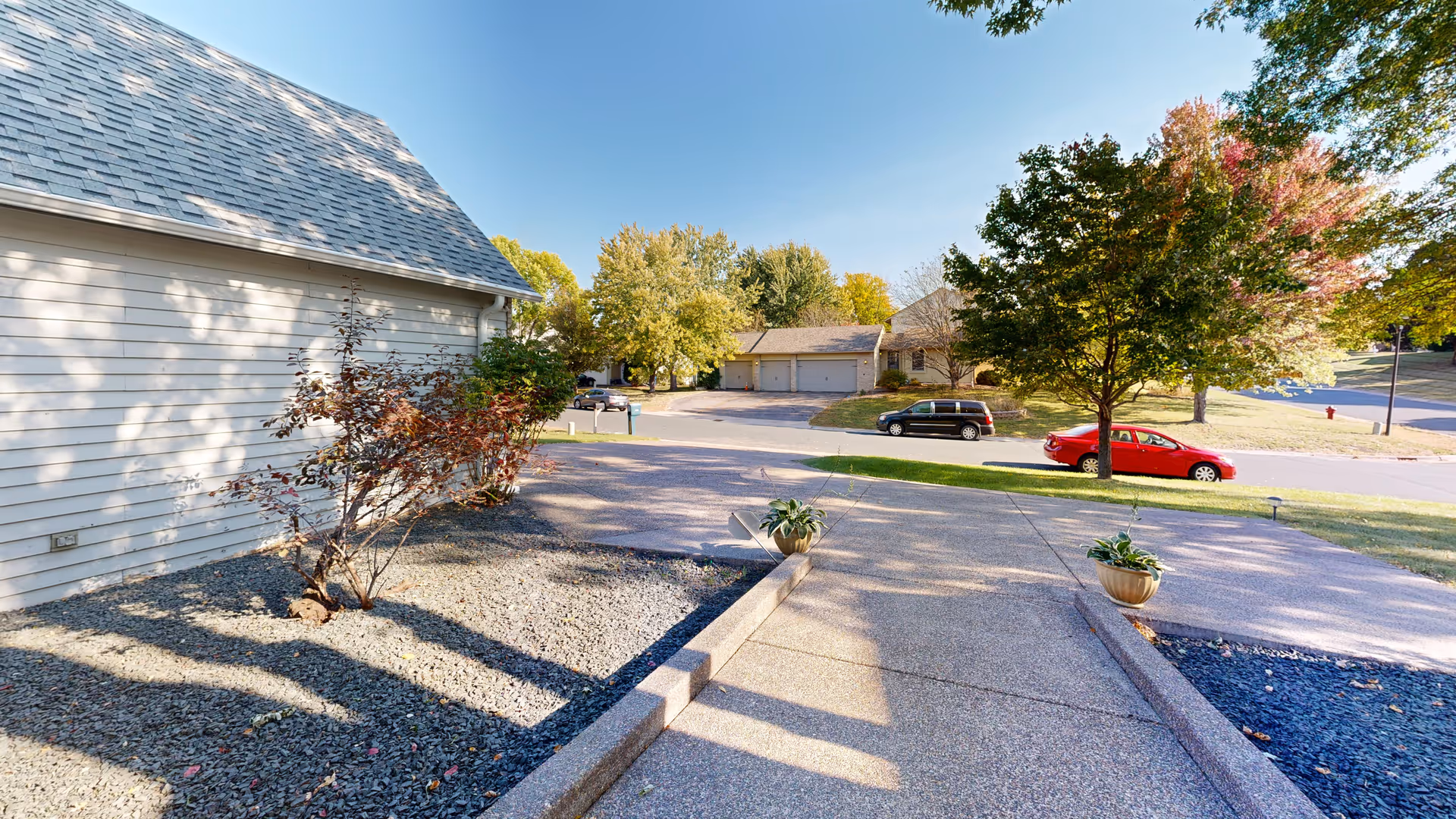 A paved driveway leading to a residential street with parked cars and trees with autumn foliage. The side of a light-colored house with a shingled roof and a small garden area with gravel and a shrub is visible on the left.