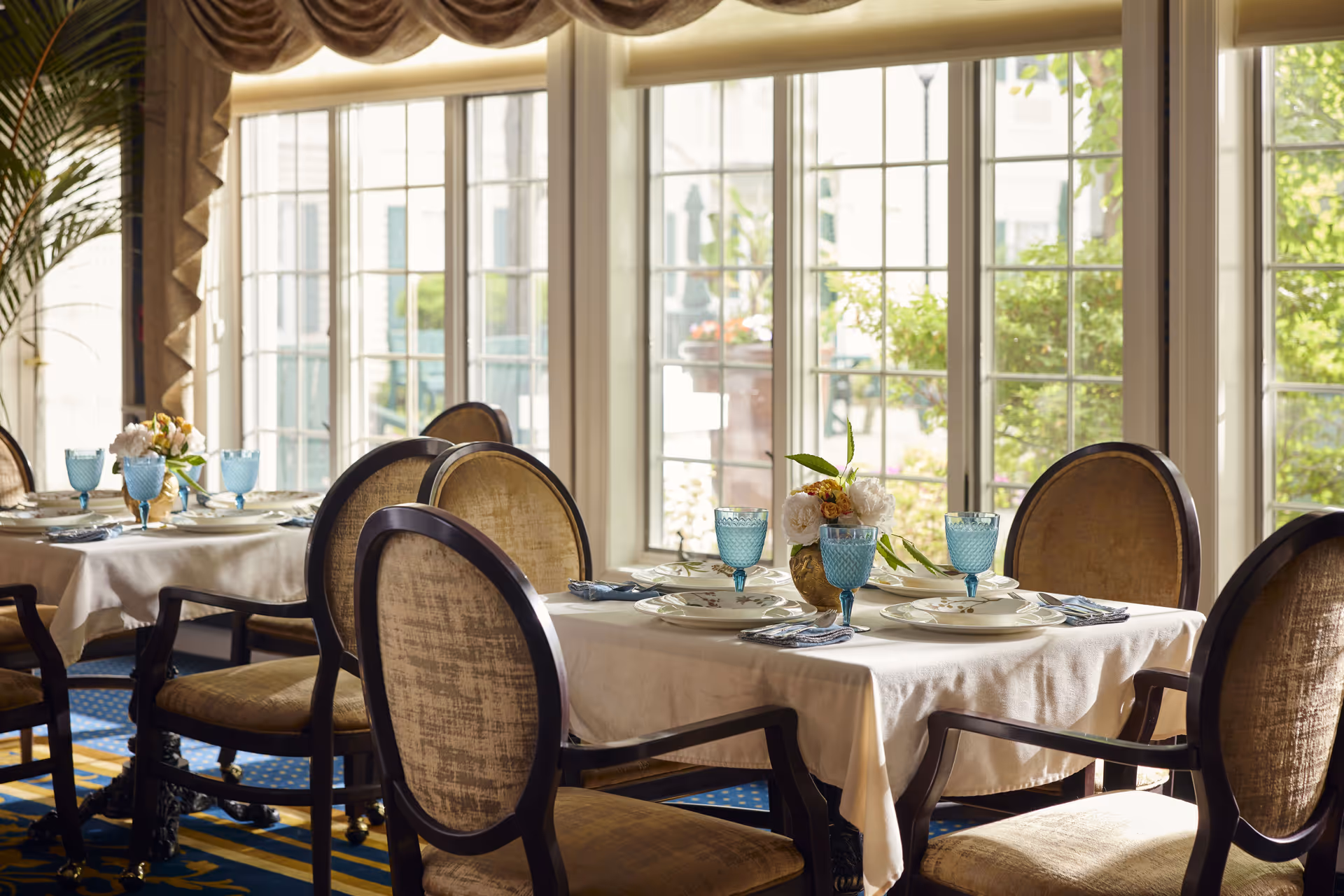 A dining room with tables covered in white tablecloths, set with plates, blue goblets, and napkins. The room has large windows letting in natural light and offering a view of greenery outside. Elegant upholstered chairs surround the tables, and a small floral centerpiece decorates each table.