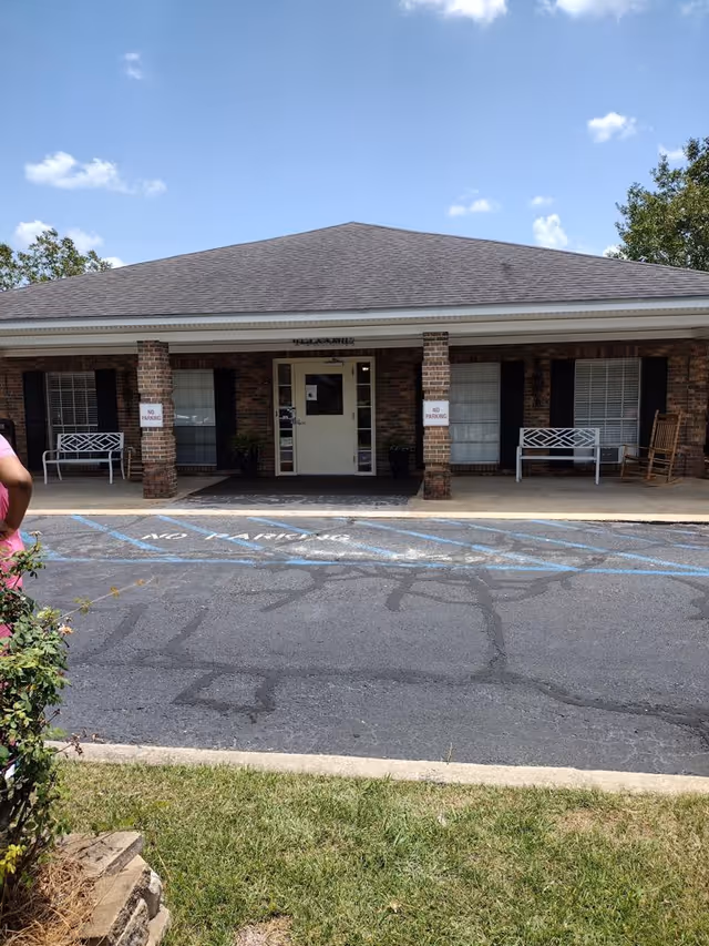 Front exterior view of a single-story brick building with a covered entrance supported by two brick columns. There are two white benches and a rocking chair on the porch. The driveway in front has 'NO PARKING' painted on it and signs on the columns also say 'NO PARKING'. The sky is partly cloudy and there is some greenery visible on the sides.