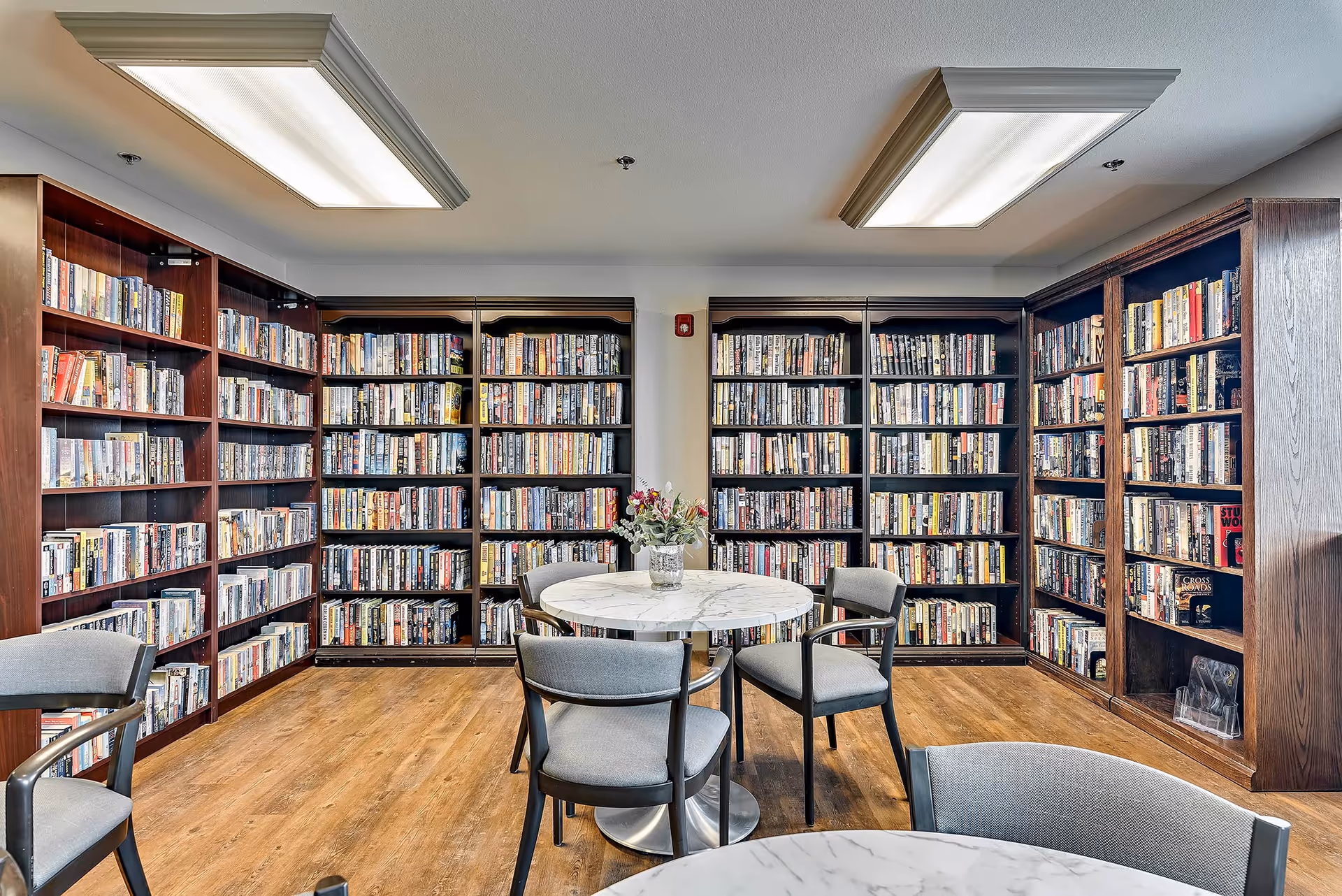 Well-lit library/reading room with bookshelves lining the walls and round marble tables with chairs in the center.