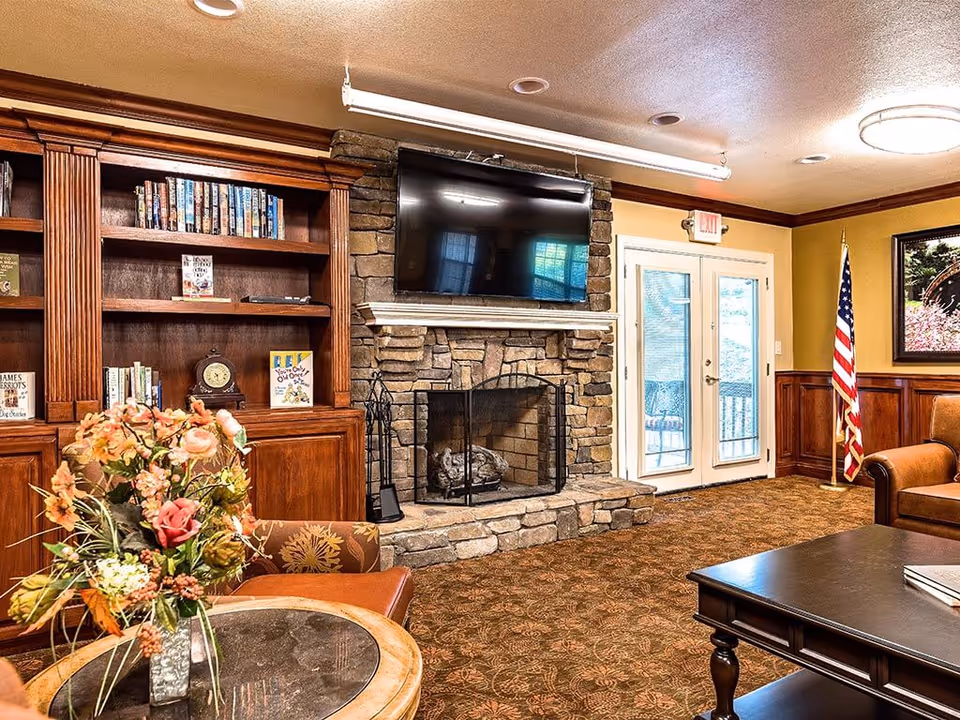 Cozy senior living common room with a stone fireplace and wall-mounted TV, bookshelves, seating, and a floral centerpiece on a table.