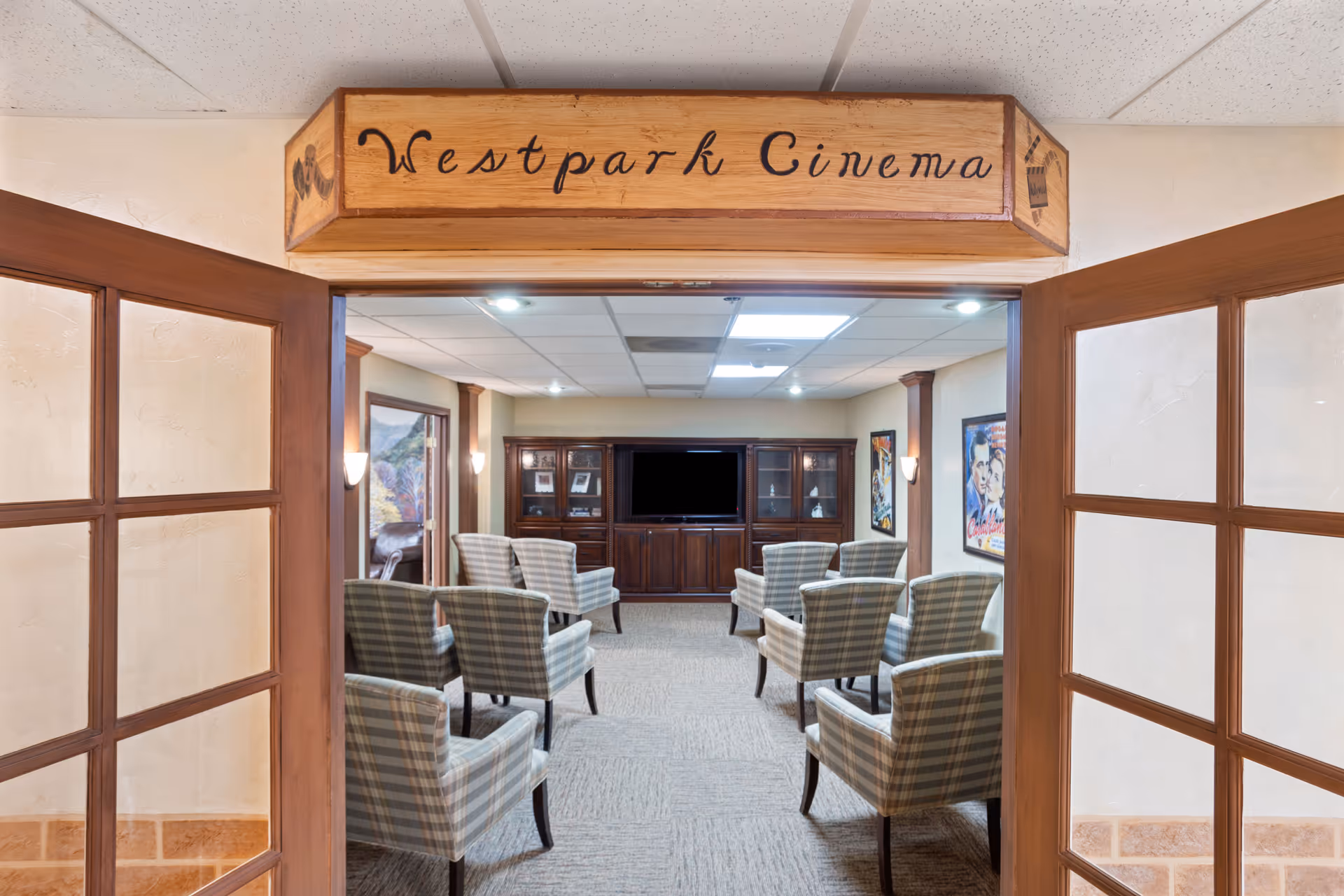View through open wooden double doors into a small cinema room named Westpark Cinema. The room contains several plaid upholstered armchairs arranged in rows facing a large flat-screen TV set in a wooden entertainment center. Movie posters are visible on the walls.