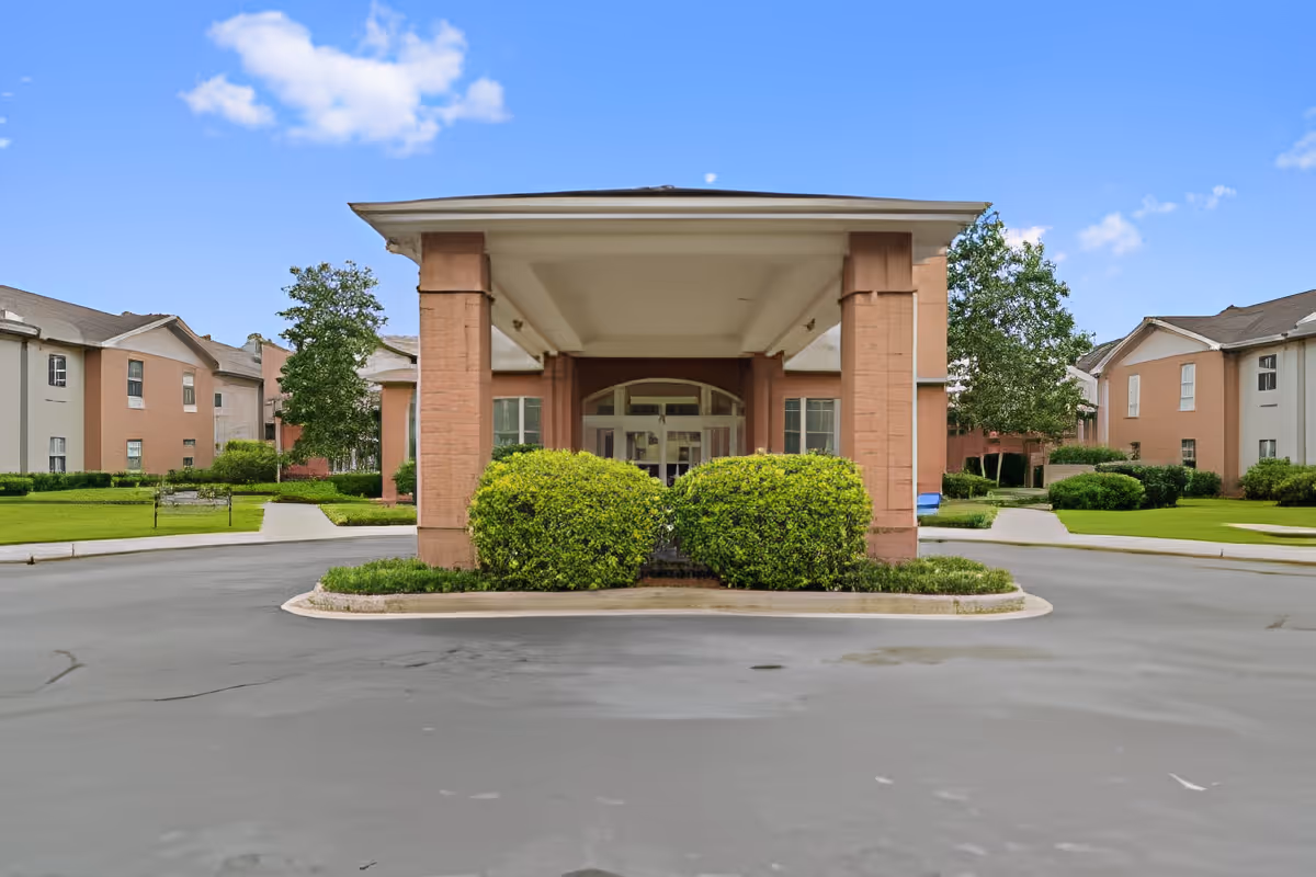 Front entrance of a senior living facility with a covered drop-off area supported by brick columns, surrounded by green bushes and well-maintained lawns. The building has multiple windows and a clear blue sky in the background.