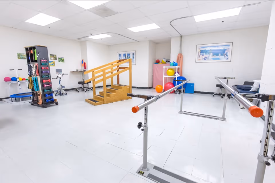A bright physical therapy room with parallel bars for walking exercises, a wooden staircase with handrails for step training, various exercise balls, weights, and fitness equipment neatly arranged around the room. The walls are white with framed pictures, and the floor is a clean white tile.