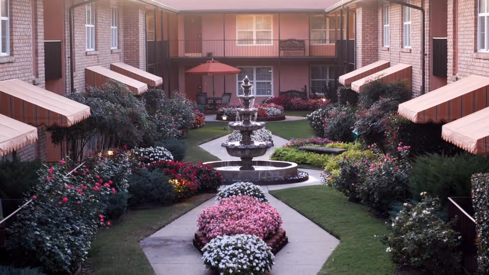Courtyard with a multi-tiered fountain, paved pathways, colorful flowerbeds, and surrounding apartment balconies with striped awnings.