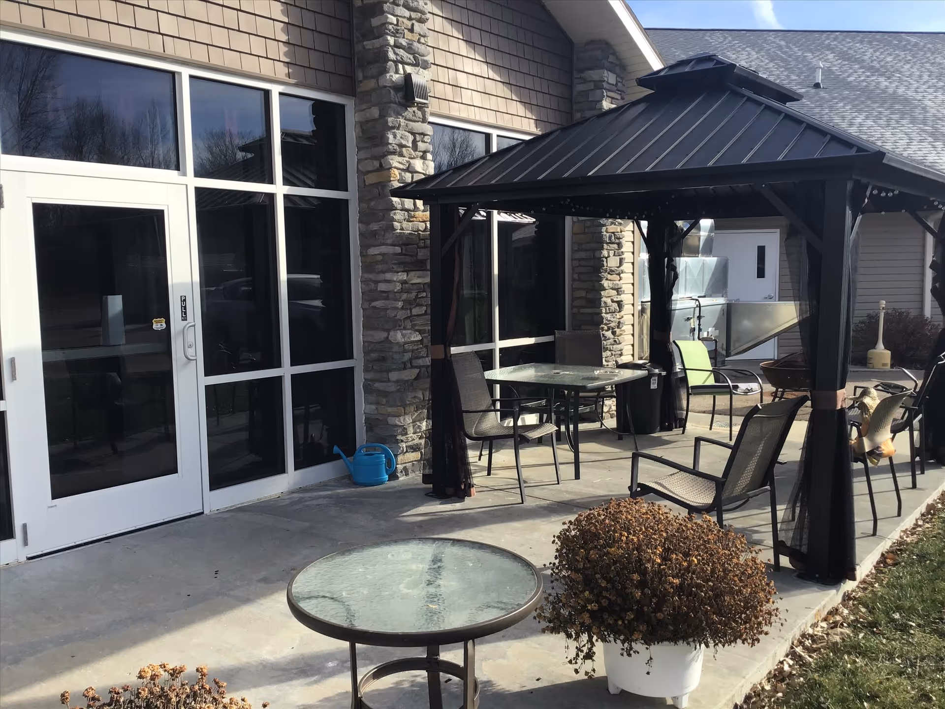 Outdoor patio with a gazebo, glass-top tables, chairs, and potted plants outside a building with large windows.
